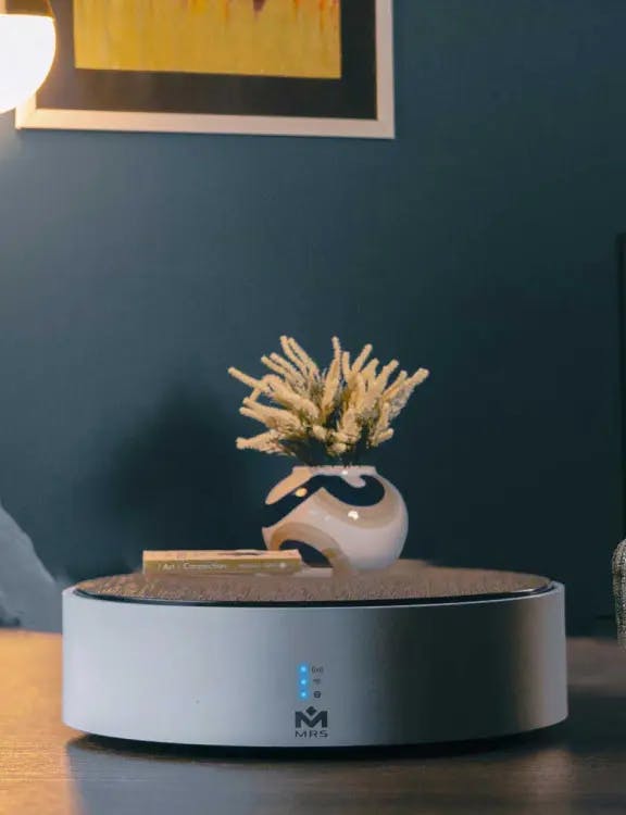 "MTronic MHub placed on a brown table with three blue vertical lights on and the MRS logo in the bottom centre; a vase, blue walls and a yellow poster with white and black outline in the background.